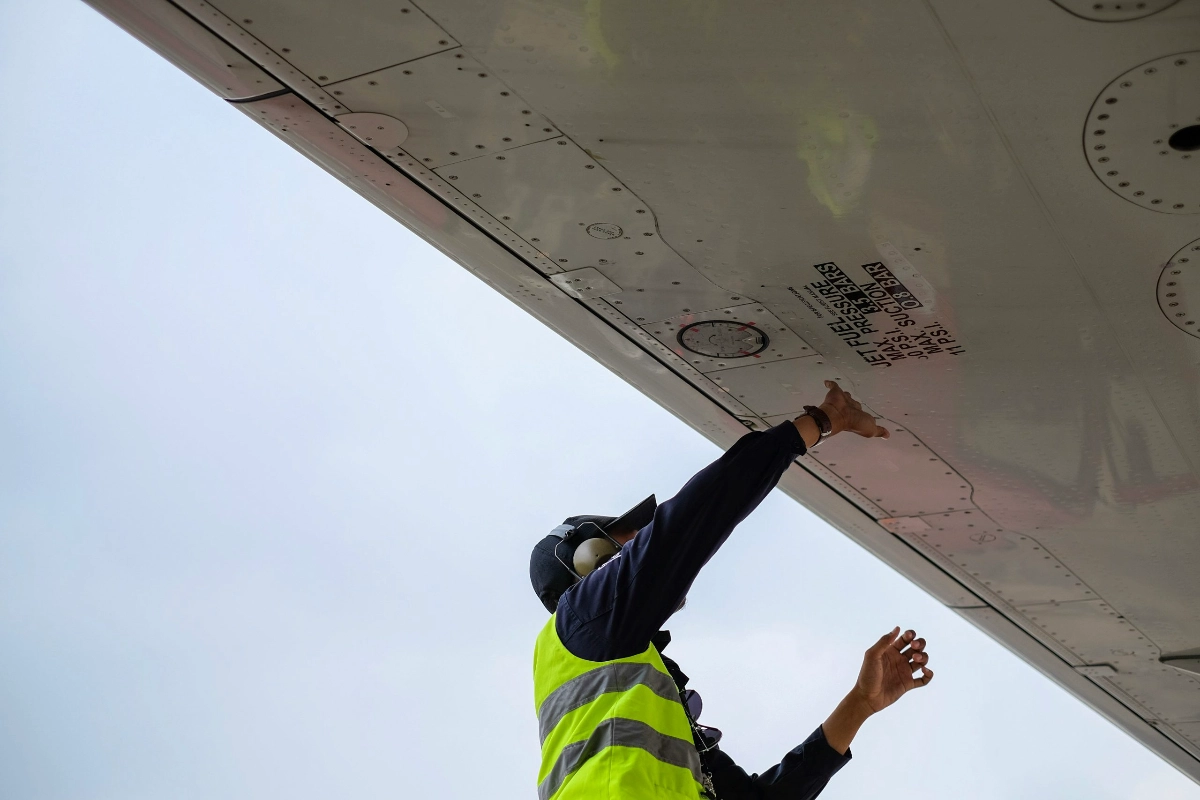 A technician in high-visibility safety gear performing a technical inspection on the undercarriage of a large aviation asset.